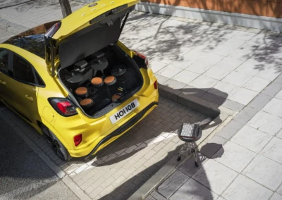 Coffre ouvert d'une Ford Puma jaune avec un kit de batterie électronique à l'intérieur, stationnée sur un trottoir pavé.