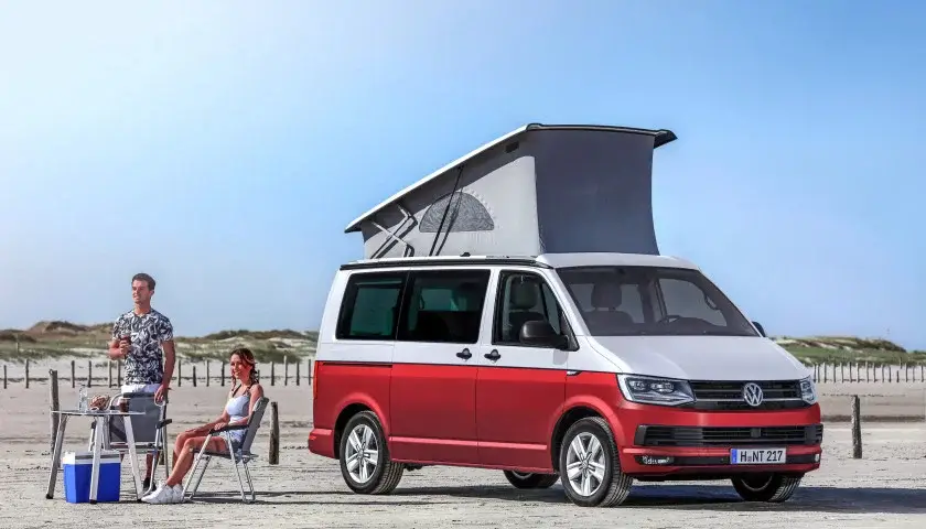 Un van Volkswagen rouge et blanc avec toit relevé est garé sur une plage, à côté d'un couple assis à une table.