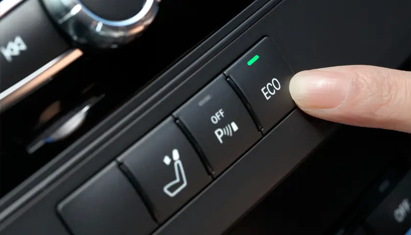 Doigt appuyant sur le bouton "ECO" du tableau de bord d'une voiture, à côté des commandes de siège et de stationnement.