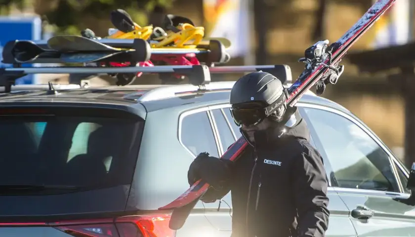 Un skieur en tenue noire porte des skis rouges à côté d'une voiture avec des skis sur le toit.