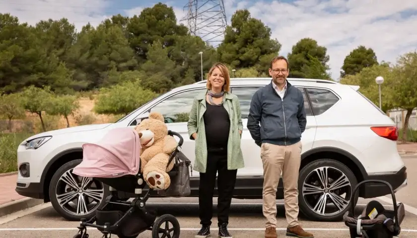Un couple devant une voiture blanche SEAT avec une poussette rose et un siège auto dans un parking arboré.
