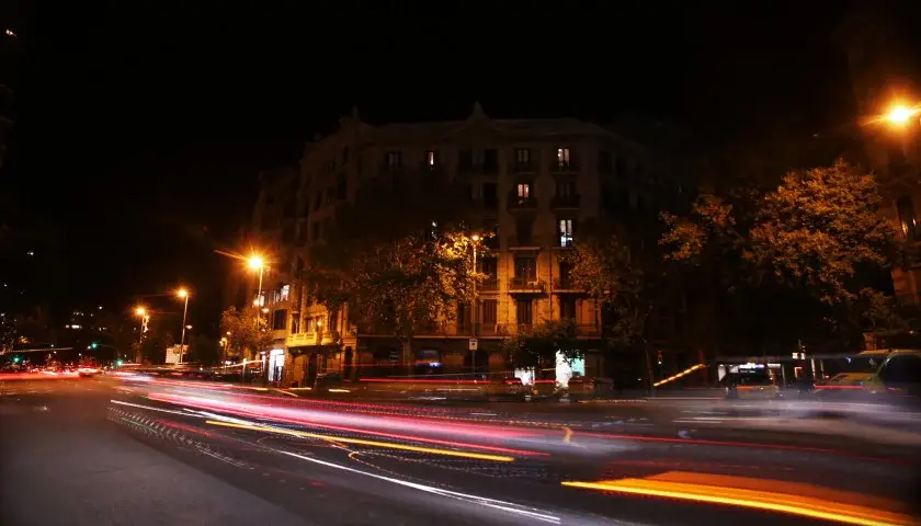 Traînées lumineuses de voitures sur une route urbaine la nuit, avec un bâtiment éclairé en arrière-plan et des arbres.