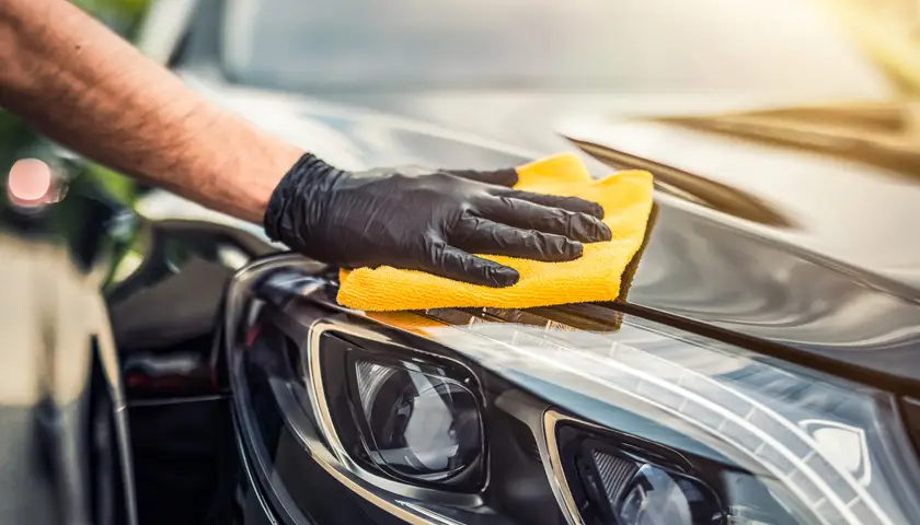 Main gantée nettoyant un phare de voiture noire avec un chiffon jaune.