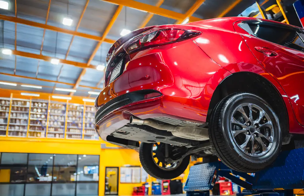 Voiture rouge sur un pont élévateur dans un atelier mécanique moderne avec éclairage au plafond et étagères en arrière-plan.
