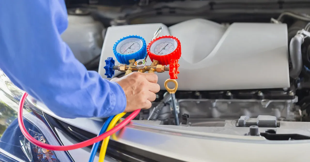 Un technicien en bleu vérifie la climatisation d'une voiture avec un manomètre à cadrans rouge et bleu sous le capot.