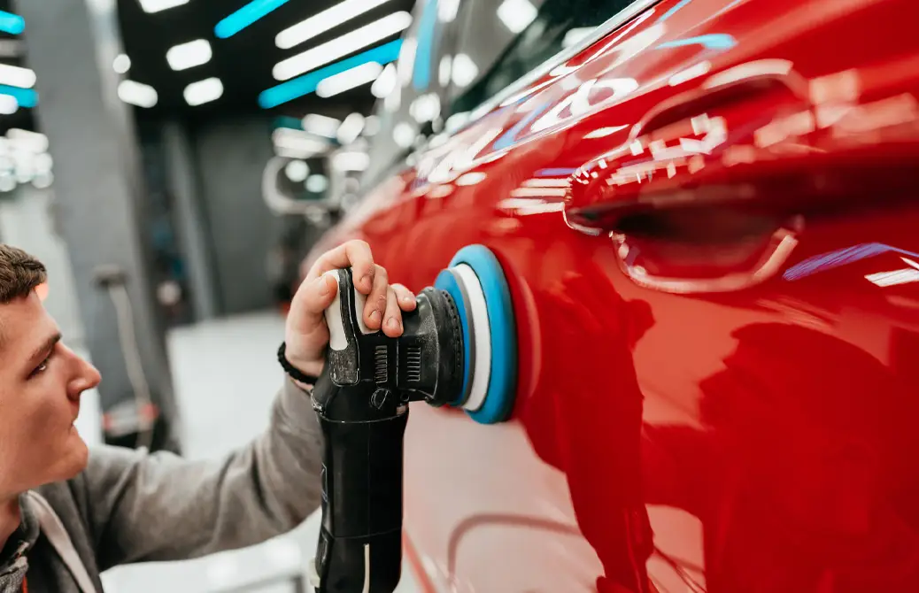 Un technicien polit la carrosserie rouge d'une voiture à l'aide d'une polisseuse électrique dans un atelier bien éclairé.
