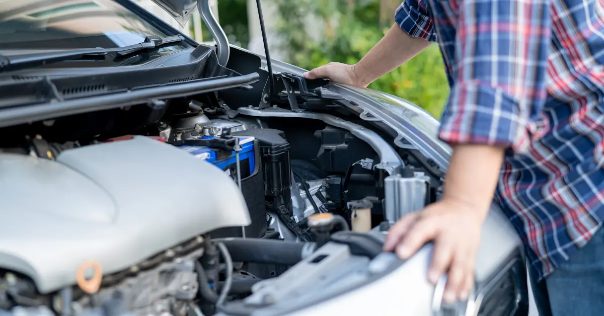 Une personne inspecte le moteur d'une voiture grise, capot ouvert, portant une chemise à carreaux.