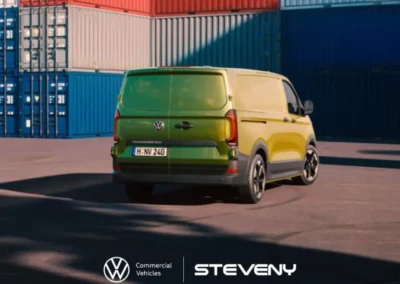 Fourgon vert Volkswagen Transporter T7 garé devant des conteneurs colorés, photo prise sur un site industriel.