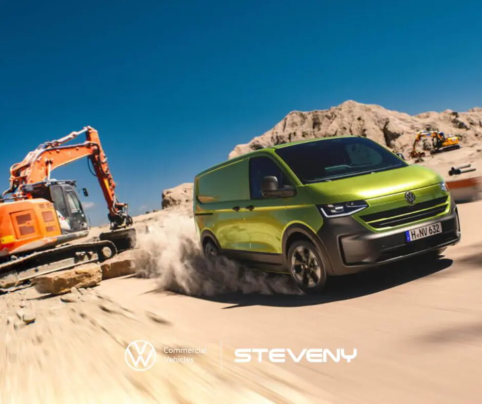 Un Volkswagen Transporter T7 vert roule sur un chantier, à côté d'une pelleteuse orange sous un ciel bleu.