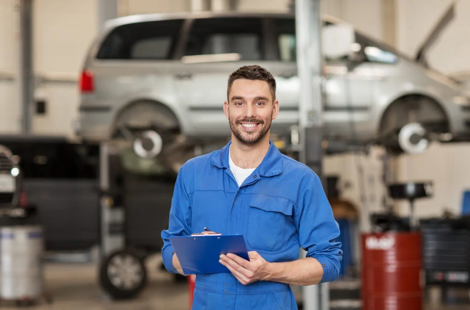 Technicien en bleu de travail tenant un clipboard, devant une voiture grise sur un pont élévateur dans un atelier.