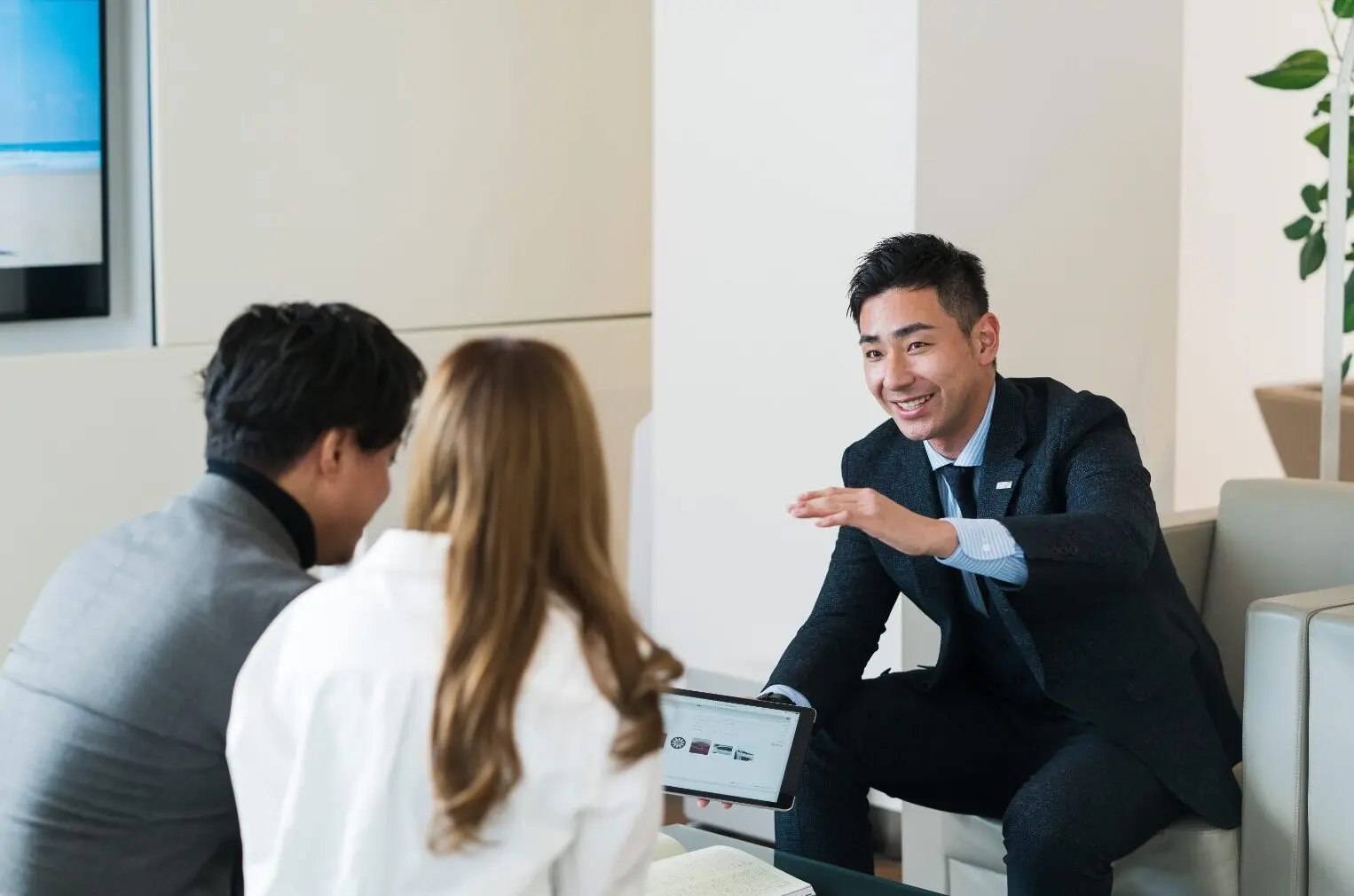 Un vendeur en costume sombre discute avec un couple dans un salon, tenant une tablette affichant des logos automobiles.