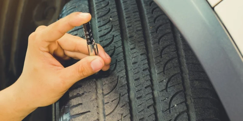 Main vérifiant la profondeur de la bande de roulement d'un pneu avec une jauge d'usure en métal sur une voiture.